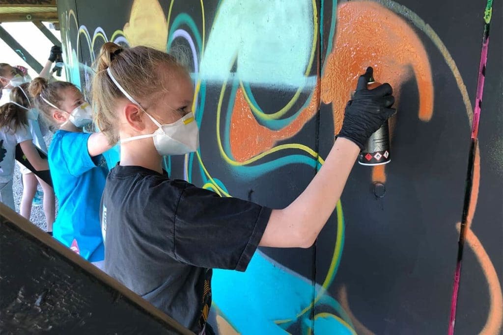 Um den Graffiti Kindergeburtstag von Salome gebührend zu feiern, haben wir von Graffiti Stuttgart mit ihren Gästen ein cooles Graffiti an der Außenwand vom Camp Feuerbach gemalt. Am Tag der Malaktion grundierte Micha die zu besprühende Fläche mit Wandfarbe, um dann die zuvor verabredeten Motive auf der Betonmauer vor zu skizzieren. Anschließend malte Salome mit ihren Freunden den Graffiti Style mit allen Buchstaben und Elementen farbig aus. Danach sprühte Micha noch die Konturen Outlines und die Lichteffekte.